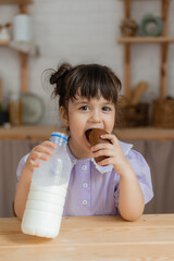 little girl in a lilac dress drinks milk and eats a bun at the kitchen table. space for text, banner