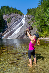 Adventurous athletic female hiker standing at the base of a waterfall in the Pacific Northwest, holding her French Bulldog up in the air.
