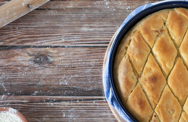 Freshly baked pie in a baking pan on a wooden background dusted with flour and a rolling pin. Top table view, copy space. Traditional homemade crusty bread, Macedonian cuisine, Balkian pastry food.