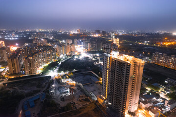 aerial drone dusk twilight shot showing orange lights of streets, homes and markets surrounding a skyscraper with the city scape stretching into the distance in gurgaon haryana delhi - obrazy, fototapety, plakaty