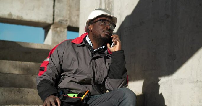 African american builder talks on smartphone. Builder sits on concrete stairs at construction site and speaks on cell phone, looks around