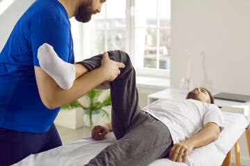 Chiropractor, osteopath or physiotherapist at modern massage room or rehabilitation center examining patient, treating pain and checking his knee flexion. Physiotherapy treatment concept