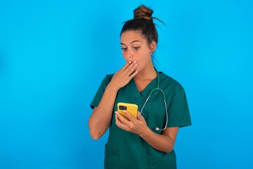 beautiful doctor woman wearing medical uniform over blue background being deeply surprised, stares...