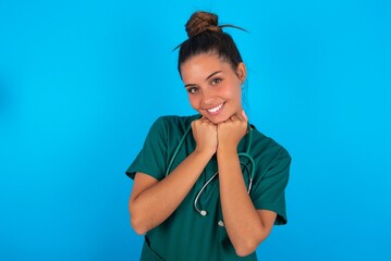 Satisfied beautiful doctor woman wearing medical uniform over blue background touches chin with both hands, smiles pleasantly, rejoices good day with lover