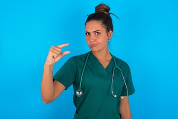 Displeased beautiful doctor woman wearing medical uniform over blue background shapes little hand sign demonstrates something not very big. Body language concept.