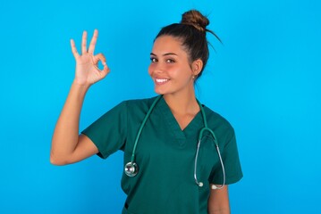 beautiful doctor woman wearing medical uniform over blue background hold hand arm okey symbol...