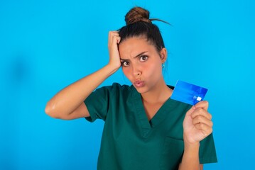 Photo of beautiful doctor woman wearing medical uniform over blue background amazed shocked hold credit card payment