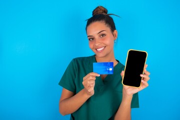 Naklejka premium Photo of adorable beautiful doctor woman wearing medical uniform over blue background holding credit card and Smartphone. Reserved for online purchases