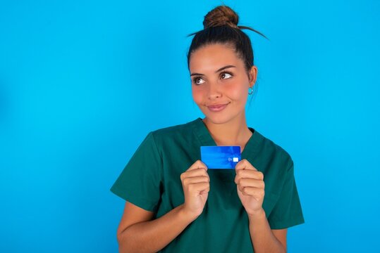 Photo of cheerful beautiful doctor woman wearing medical uniform over blue background hold debit card look empty space