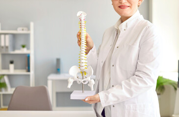 Doctor at orthopedic clinic showing anatomical spine model. Cropped shot of happy woman in white lab coat holding model of human backbone. Medicine, posture, bone health concept. Medical background