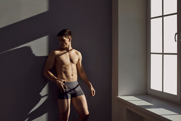 Sexy man with muscular body in black classic underpants posing by window in sunlight. Man with...