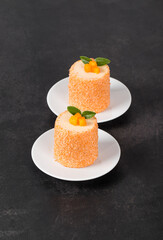 Apricot mousse cream chocolate dessert of cylindrical shape, sprinkled with colored coconut flakes, on a plate. Dark background
