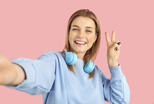 Happy cheerful confident young online social media blogger girl takes selfie photo on pink color background. Positive high school student holding mobile phone, smiling and doing peace victory gesture