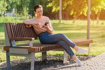 Beautiful brunette with a mobile phone sits on a bench in the park - Powered by Adobe