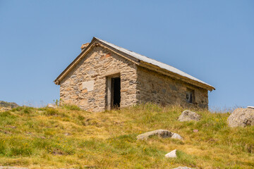 Stone mountain shelter at an altitude of 2000 meters, without a door, on a green grass meadow with some scattered stones, with direct sunlight and a blue sky without any clouds.