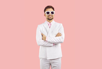 Portrait of funny cool and stylish young man in suit isolated on pastel pink background. Confident modern businessman in white suit and sunglasses posing with folded arms and smiling. Banner.