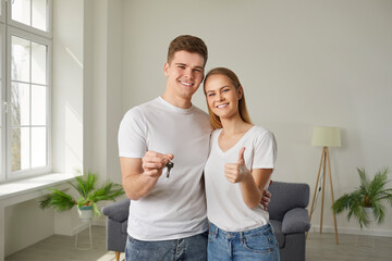 Happy beautiful newlywed couple buys a house. Portrait of a cheerful young family showing the key to their new home, smiling and giving a thumbs up. Real estate, mortgage, purchasing property concept