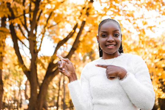 Smiling laughing african american woman standing in the city park, having fun, point fingers on copy space for text or picture. Autumn season, yellow red and orange leaves. Fall time.
