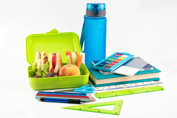 On a white background, a lunch box with a sandwich, vegetables and fruits, a bottle of water and school supplies.