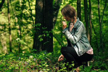 a young beautiful woman in casual clothes walks through the green forest in the warm season and admires the lilac small forest flowers. Leisure active young people in nature