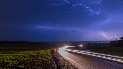 Fototapeta premium Gewitterblitze über einer Strasse, wo Autoscheinwerfer Leuchtspuren hinterlassen, Langzeitbelichtung