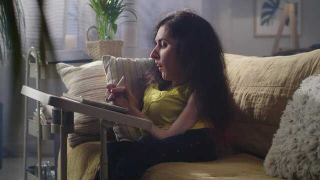 Concentrated woman with spinal muscular atrophy sitting on a couch at cozy home and drawing on digital tablet computer using pencil