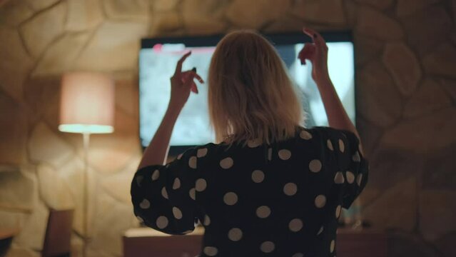 Woman dances to music on big TV raising hands. Blonde lady tourist enjoys dancing and relaxing on vacation in luxury hotel suite closeup