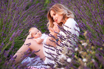 Happy mother with pretty daughter on lavender background. Beautiful woman and cute baby sitting in meadow field. Family holiday in summer day.