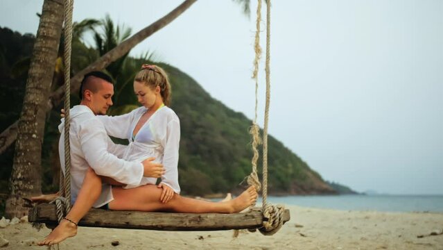 Cute loving couple on the swing on tropical beach. Woman and man in white shirt enjoy gold sunset and life in beaches, palm tree. Evening warm sunset. Summer holiday vacation tropical tourism concept