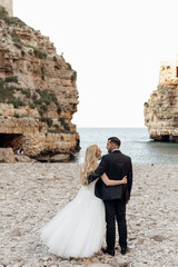 Back view of wedding couple standing on stony shore of Adriatic sea near magnificent rocks, meeting eyes. Vertical.
