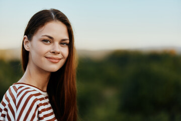 Close-up portrait of a young woman with a beautiful smile looks at the camera in a striped t-shirt on a background of trees