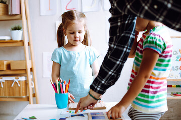 Two cute little girls pupils standing at white table, playing board games in bright classroom. Teacher helping children.