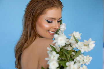 Fototapeta premium Photo closeup of lovely smiling woman with bright evening makeup and highlighting cheekbones on blue background, white flower decoration. Advertising shooting, professional makeup and beauty products