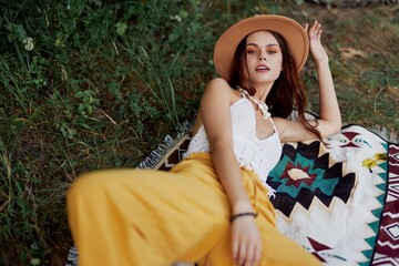 A woman in eco-friendly clothing in a hippie look is lying on a colored plaid smiling and looking at an autumn sunset in nature in the park. Lifestyle on a journey of harmony with the world