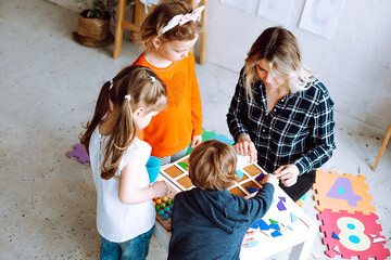 Top view of group of preschoolers and teacher. Young woman showing how to play board games collecting geometric figures.