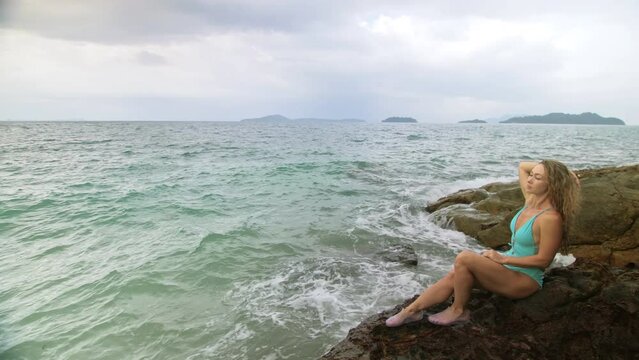 Handsome long-haired woman chill relax resting on rock of sea reef stone, stormy cloudy ocean. Woman in blue swimsuit dress tunic. Concept rest tropical resort coastline traveling outdoor tourism