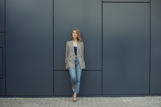 Full length photo of thinking young businesswoman standing on one leg leaning on gray wall of office building and looking up. Urban lifestyle, entrepreneur, well dressed lady, successful people