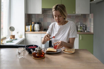 Pensive single middle-aged woman makes late breakfast sitting at table. Blonde female makes tea and sandwich spreads jam on toast sitting alone in stylish kitchen. Lady eats healthy food in morning