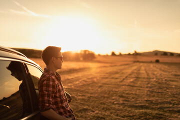 Young man relaxing on car looking out at the beautiful nature sunrise view. Travel, feelings of happiness and new day concept. 