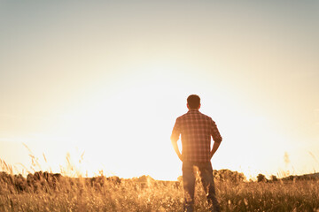 Young thoughtful man standing in a field facing the sunset. People finding peace, and happiness in nature concept. 