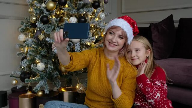 Happy mother and daughter in sweaters and red hat takes a selfie on their smart phone at christmas tree at home.