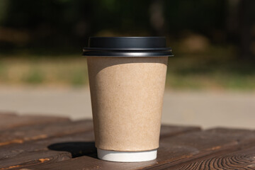 A paper cup of coffee with a lid stands on a rustic bench table against the backdrop of green...