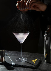 Martini glass with pale pink cocktail on the table with lemon garnish, shaker and spoon, black background