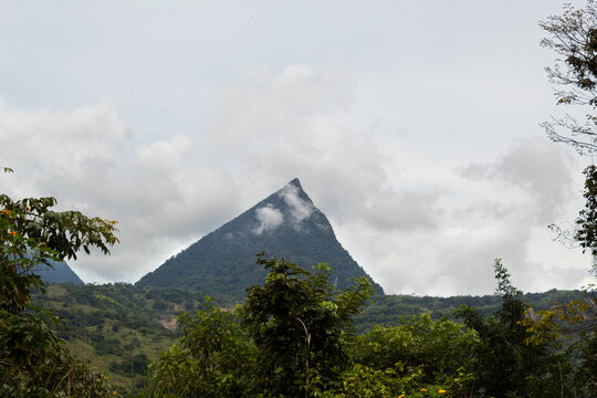 Pyramid shaped mountain with clouds in front and behind with lots of vegetation in the foreground. Cerro Tusa, Antioquia, Colombia