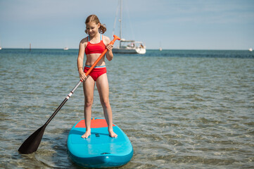 Beautiful teenage girl standing up on paddle board on blue sea at summer vacation