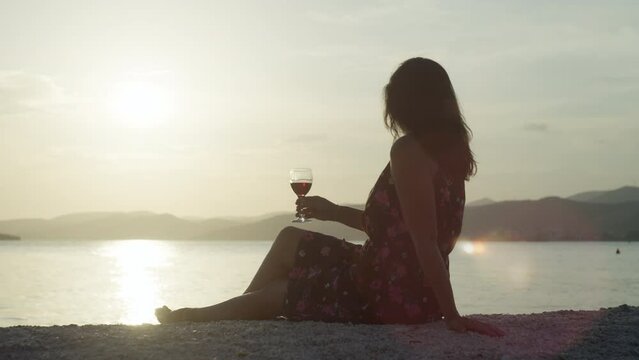Romantic landscape of the sea, a girl drinks wine on the beach at sunset. A beautiful woman with a glass in her hands looks at the nature of the ocean. High quality 4k footage