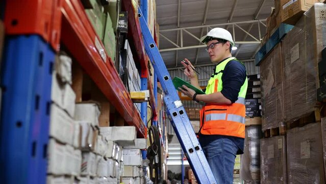 Working on ladder stair. Portrait handsome Asian male warehouse professional worker in safety vest and hardhat helmet in warehouse factory industrial. product in cardboard boxes on stored shelves.