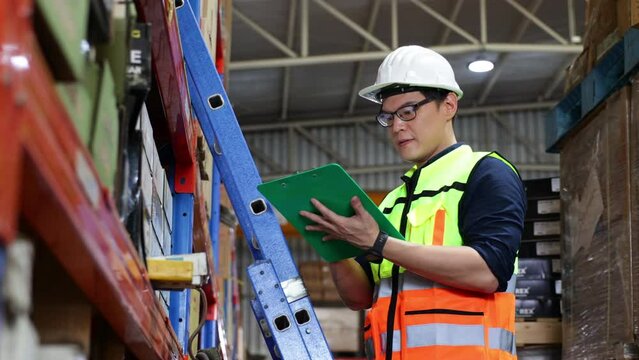 Working on ladder stair. Portrait handsome Asian male warehouse professional worker in safety vest and hardhat helmet in warehouse factory industrial. product in cardboard boxes on stored shelves.