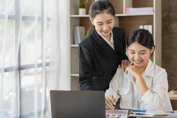 Two young Asian female accountants discuss working on investment projects and planning strategies using laptop computers in their office.