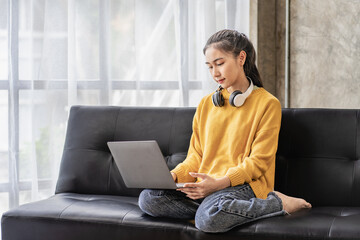 Portrait of smiling Asian woman wearing wireless headphones using laptop at home on sofa. in the living room